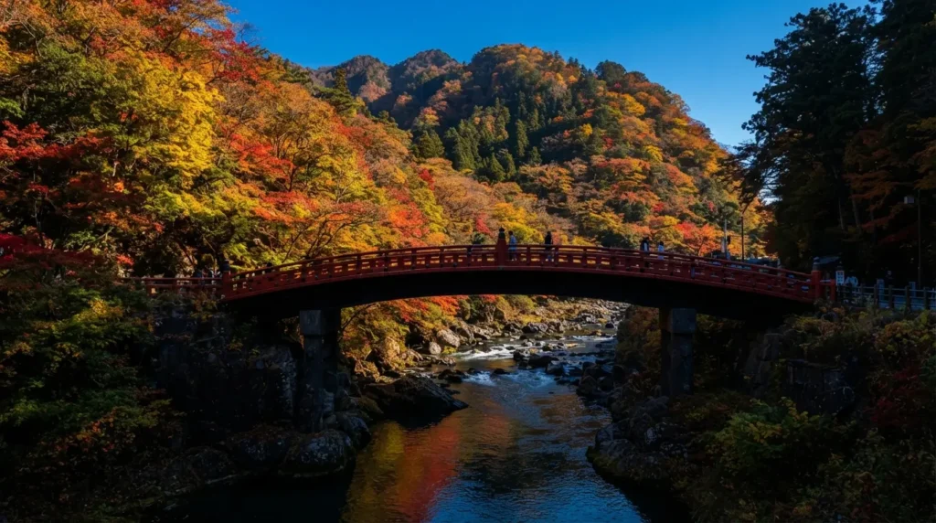 The vermilion Shinkyo sacred bridge at Nikko with vivid red and orange autumn maple trees on the mountain slopes above, the Daiya River reflecting the colours below